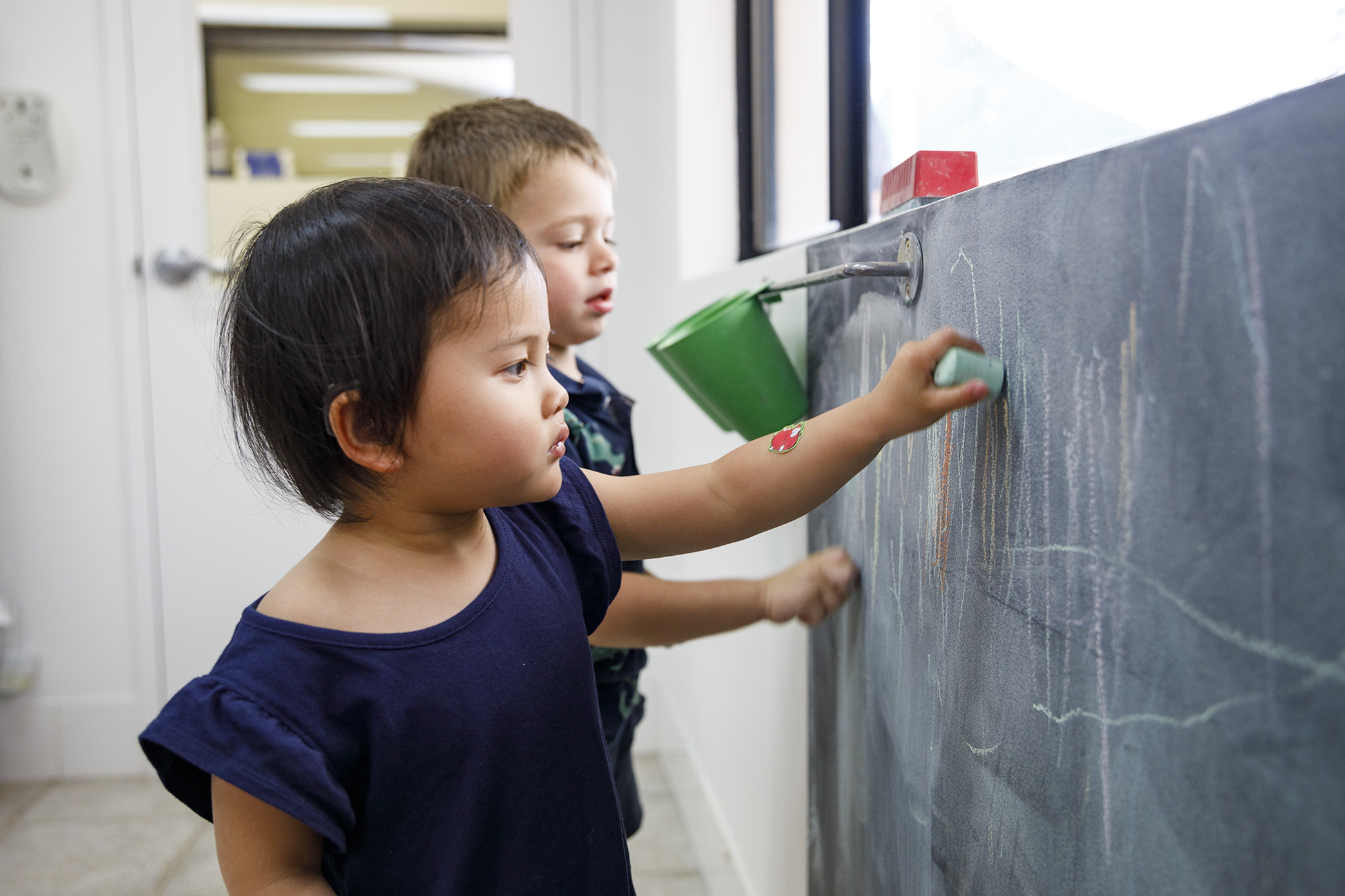 Two young children drawing with chalk on a black chalkboard.