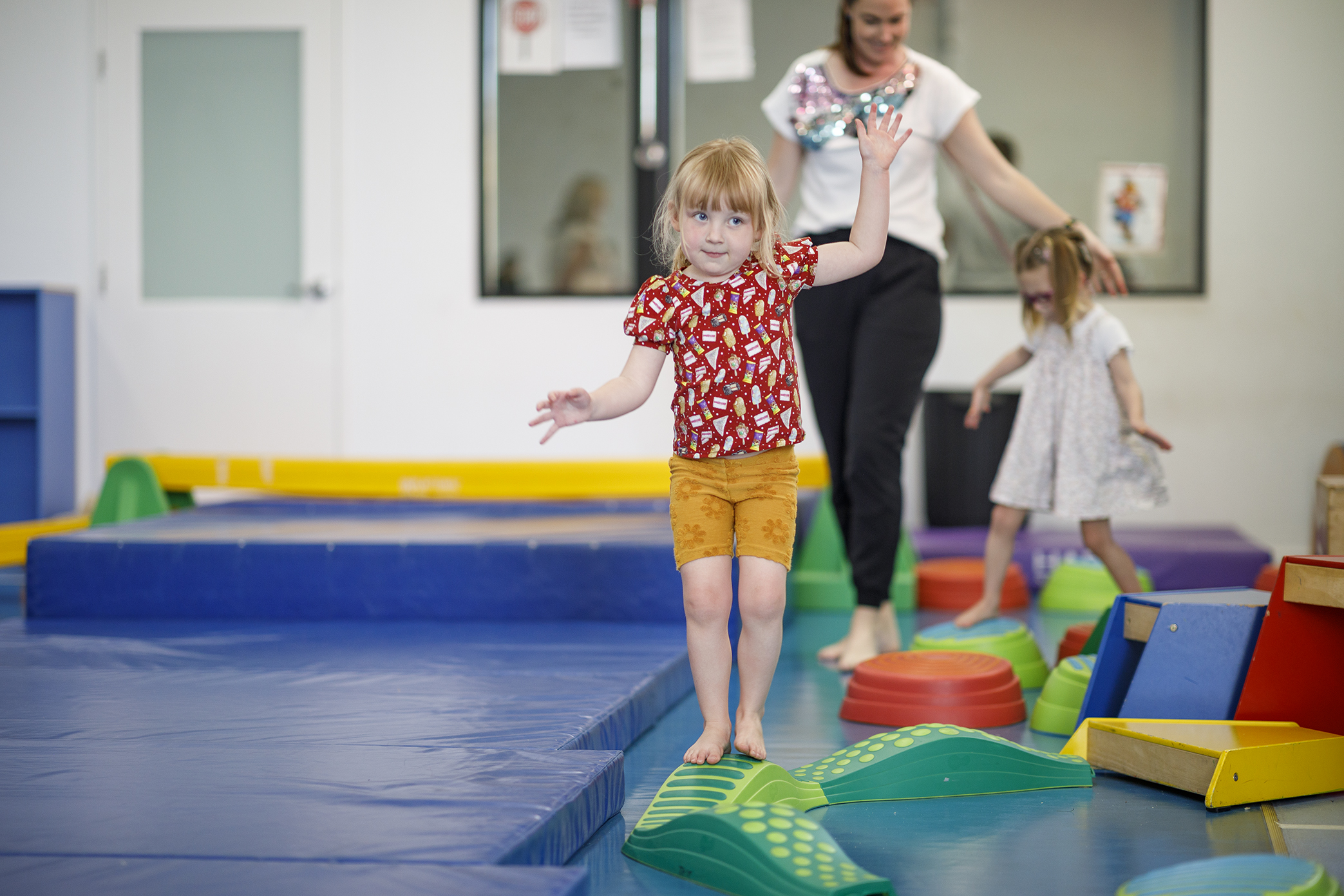 Young child in a red top and yellow shorts balancing on a low balance beam in a gymnasium. An occupational therapist and another child are in the background, also on the balance beam.