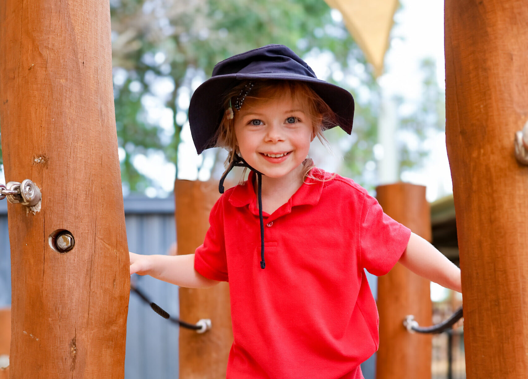 Young child on a playground smiling to the camera. Wearing a red shirt.